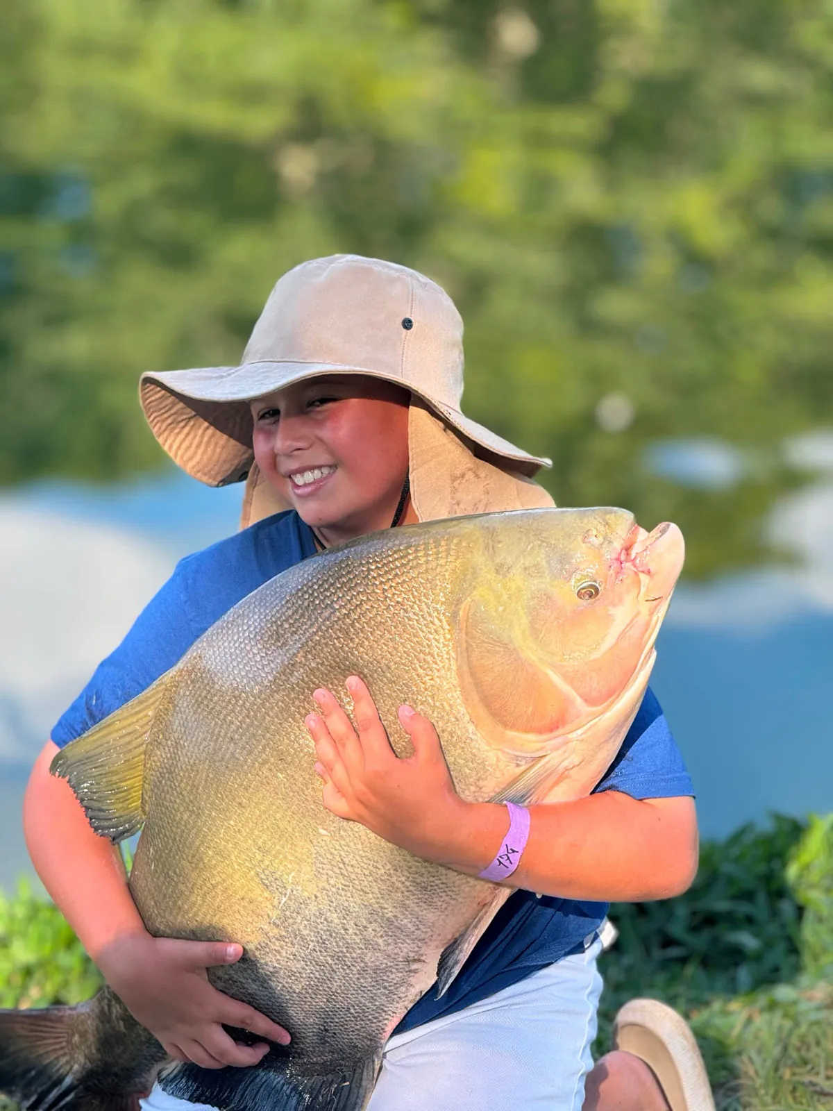 Menino de chapéu sorrindo segurando um peixe grande na pescaria, foto com celular